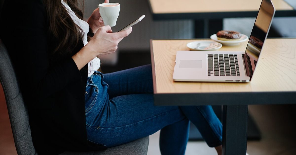 Person using a smartphone beside a laptop in a cafe