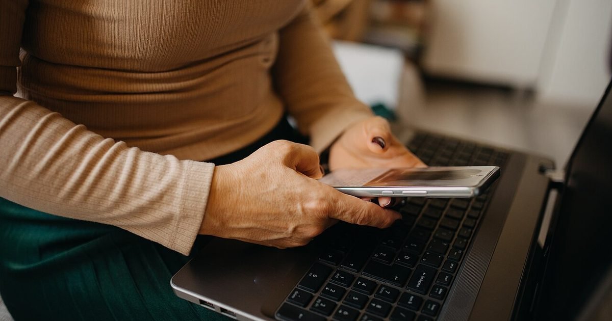 Older person holding a smartphone beside a laptop while adjusting phone settings
